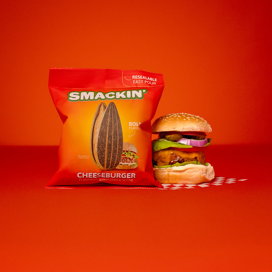 A red and yellow Smackin' Cheeseburger flavored sunflower seeds bag and a detailed cheeseburger are displayed on a vibrant red background, the bag features a large sunflower seed and "Resealable Easy Pour" text while the burger includes melted cheese fresh lettuce red onion and pickles resting on a white and red checkered pattern