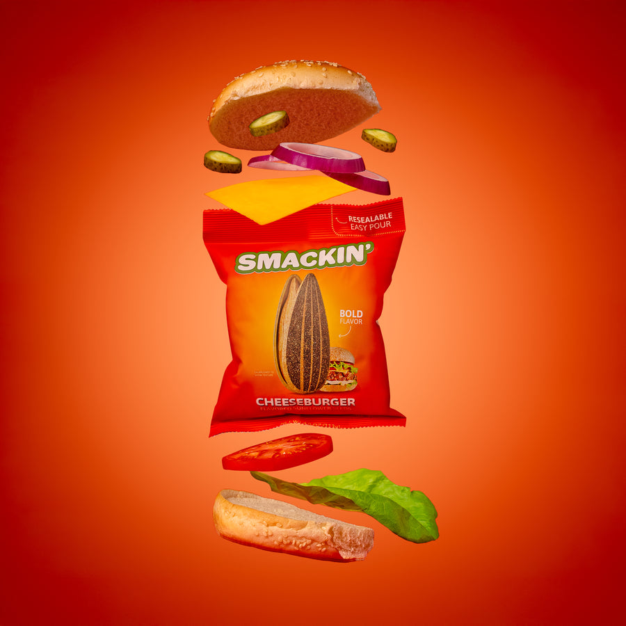 Photography, deconstructed cheeseburger ingredients like a sesame bun, cheese, red onion, pickles, tomato, and lettuce floating around a vibrant red Smackin' Cheeseburger Flavored Sunflower Seeds bag on a gradient red-orange background