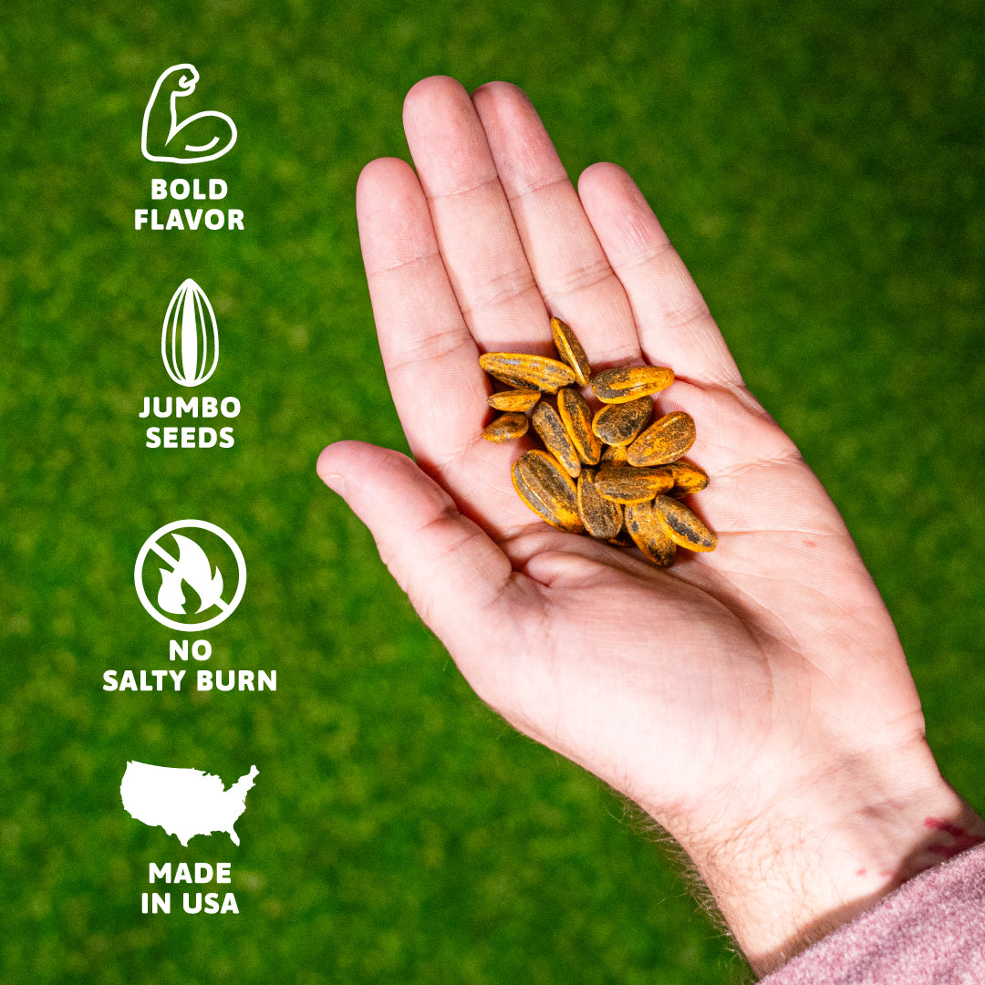 A hand presents a vibrant pile of orange-yellow jumbo sunflower seeds coated with seasoning, set against a bright green grass background with white infographic labels for Bold Flavor, Jumbo Seeds, No Salty Burn, and Made in USA