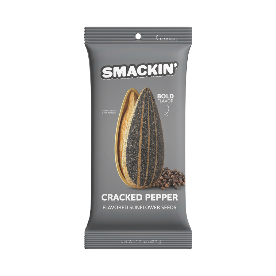 Product photography of a gray Smackin' Cracked Pepper Flavored Sunflower Seeds pouch featuring an enlarged cracked sunflower seed revealing a vibrant yellow interior and a black speckled shell, accompanied by a small pile of whole black peppercorns