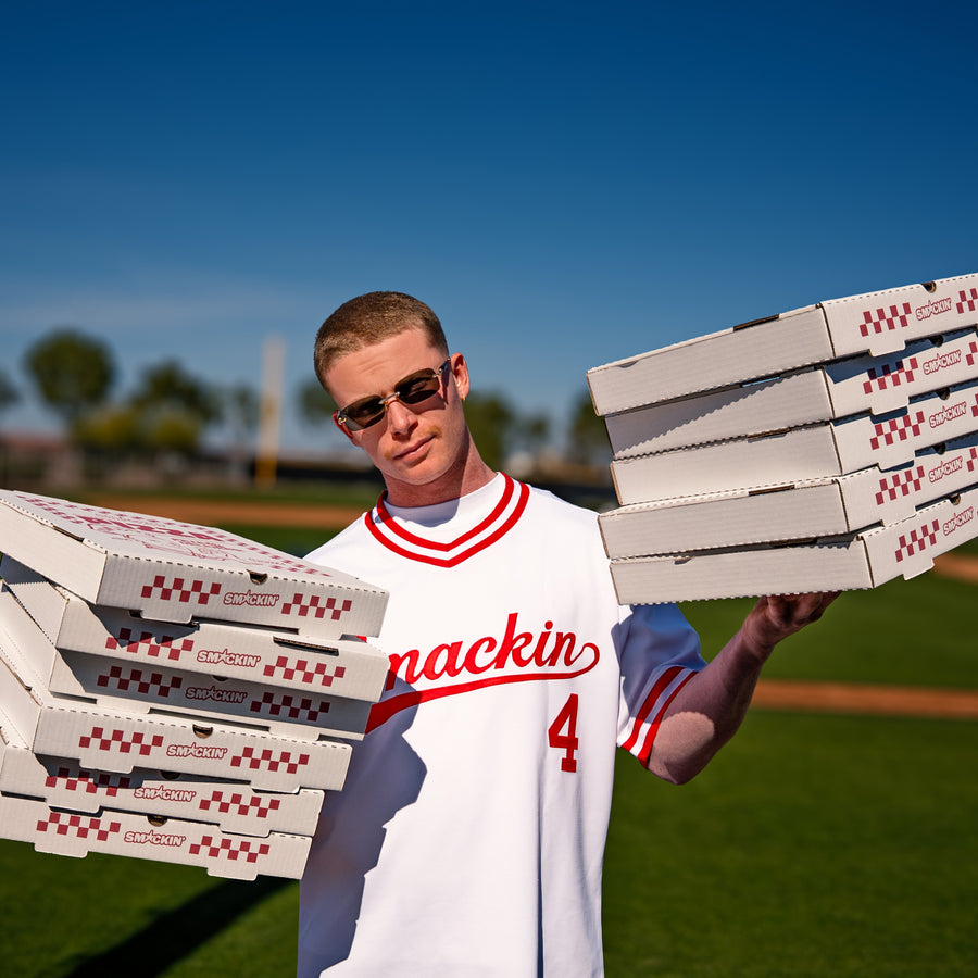 Photograph of a man in gold-rimmed sunglasses and a white baseball jersey with red trim and Smackin 4 branding holding two large stacks of Smackin pizza boxes with red checkered patterns on a vibrant green baseball field under a clear blue sky