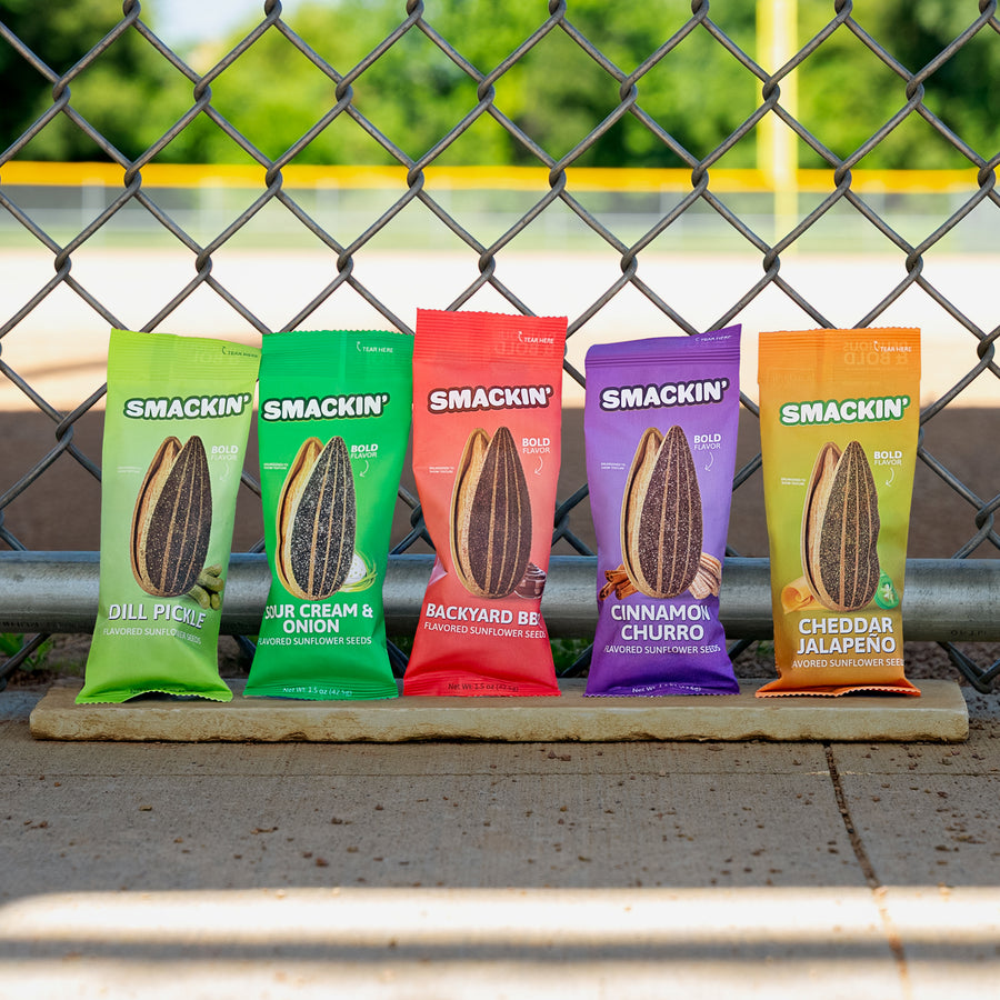 Commercial photograph of five colorful Smackin' sunflower seed bags, each with a large seed graphic and bold flavors like Dill Pickle and Cinnamon Churro, arranged on a ledge against a chain-link fence at a baseball field with a yellow line
