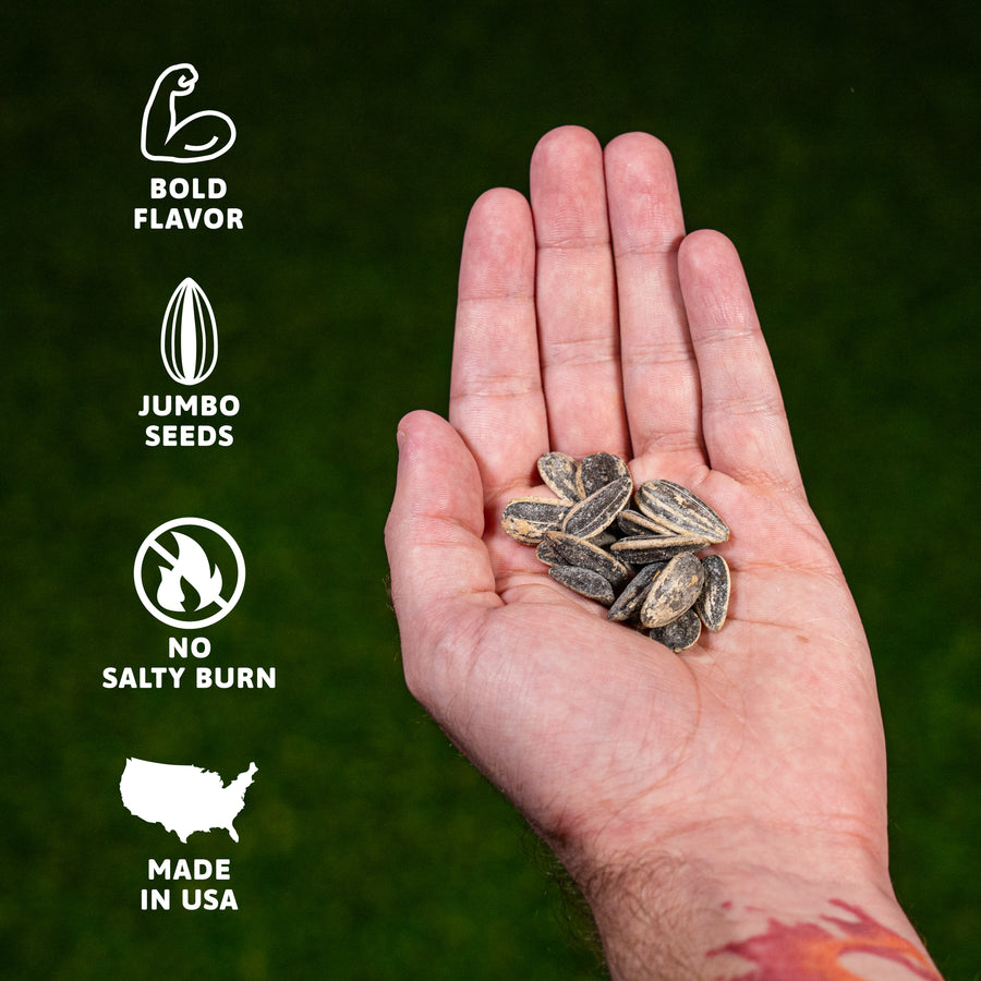 Photograph depicting a person's hand cupping numerous dark roasted sunflower seeds against a textured dark green background, with white textual overlays and icons on the left side reading 'Bold Flavor,' 'Jumbo Seeds,' 'No Salty Burn,' and 'Made in USA,' and a hint of a red and orange design on the wrist