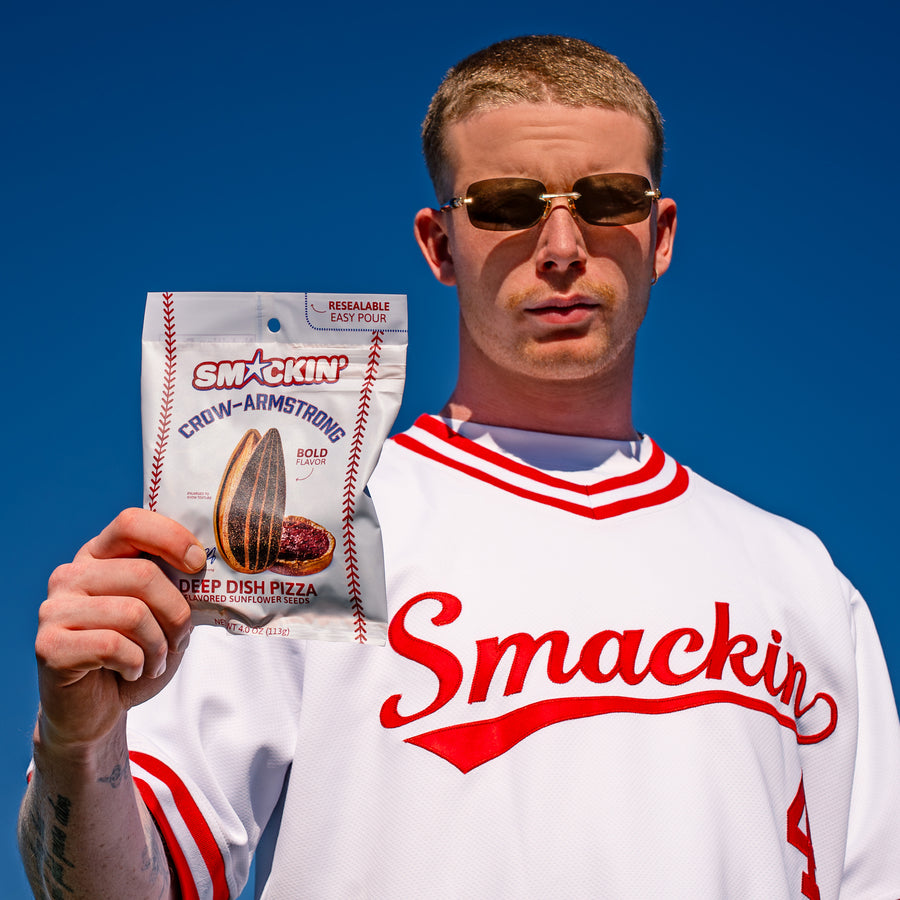 Photograph of a man in a white and red Smackin baseball jersey with gold sunglasses and a mustache holding a bag of Smackin Crow-Armstrong Deep Dish Pizza sunflower seeds, featuring baseball stitching graphics, against a vivid blue sky